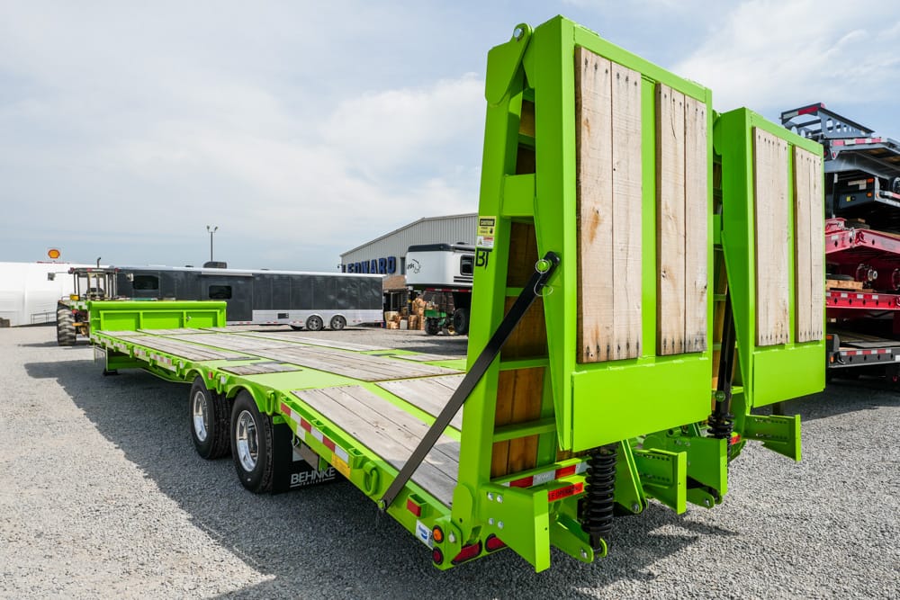 Bright green heavy-duty trailer with folding ramps and wooden deck, parked in a gravel yard under a partly cloudy sky.