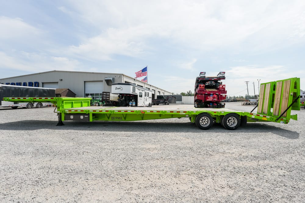 Bright green lowboy trailer with a wood loading ramp sits on a gravel lot, industrial buildings and an American flag in the background.