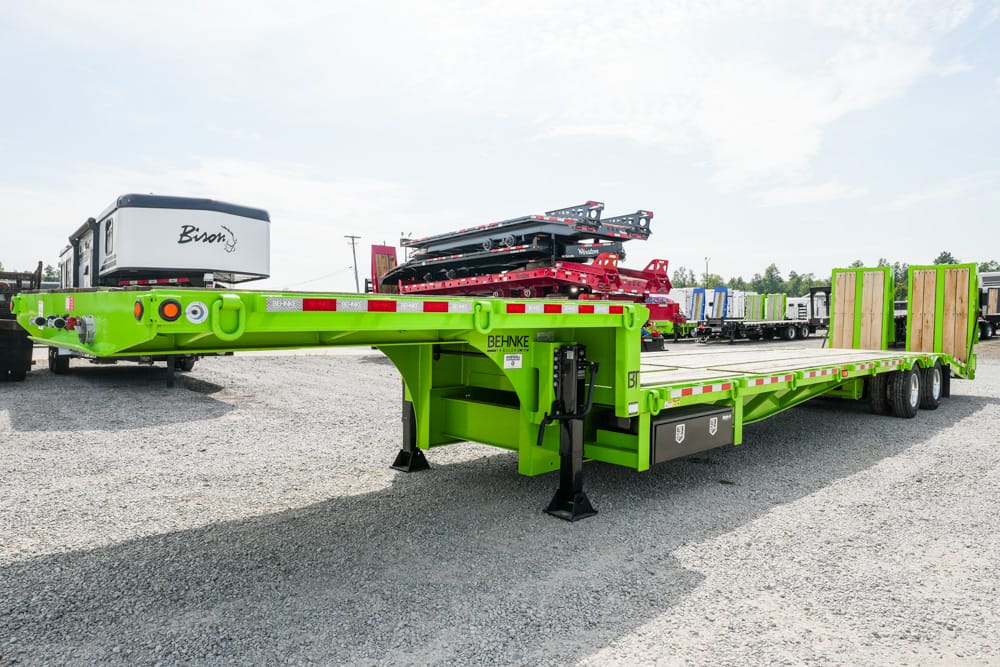Bright green lowboy trailer on a gravel lot, with rear ramps and hydraulic legs deployed.