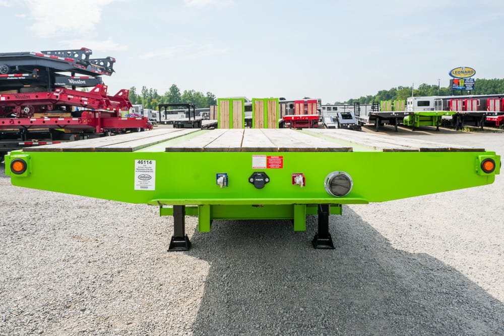Green flatbed trailer viewed from the back with a wooden deck, in a lot of trailers and equipment.