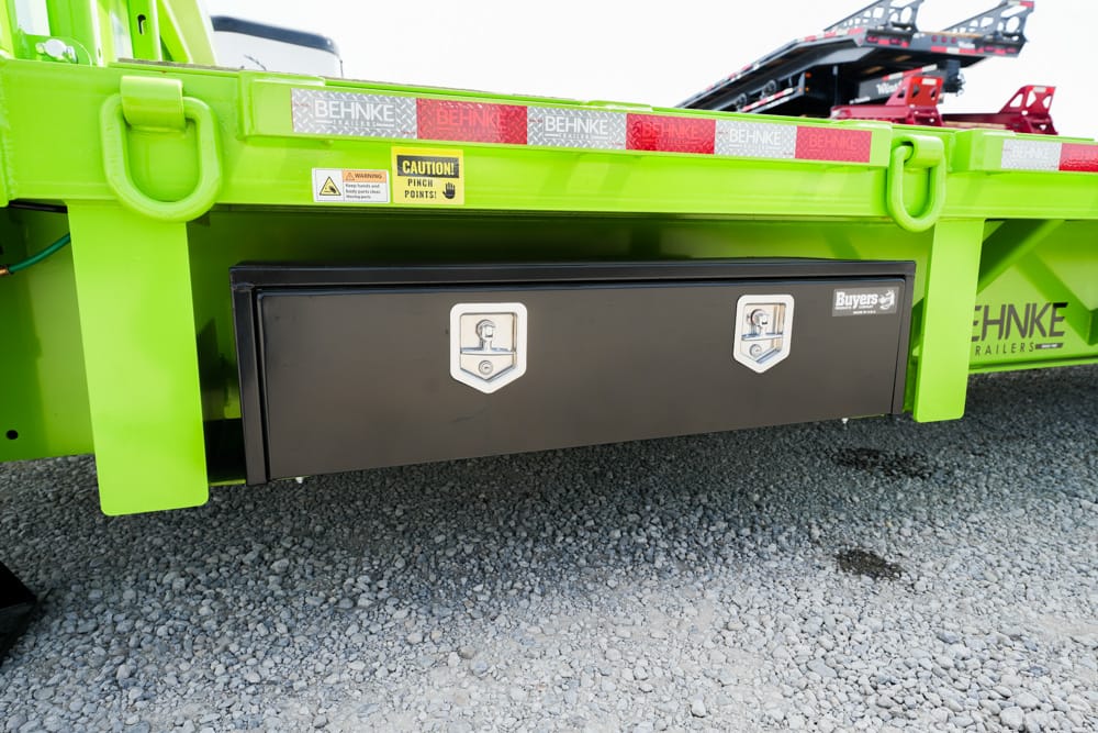 Bright lime-green trailer underside with a black rectangular storage box and two silver latches on the front panel; gravel ground beneath.