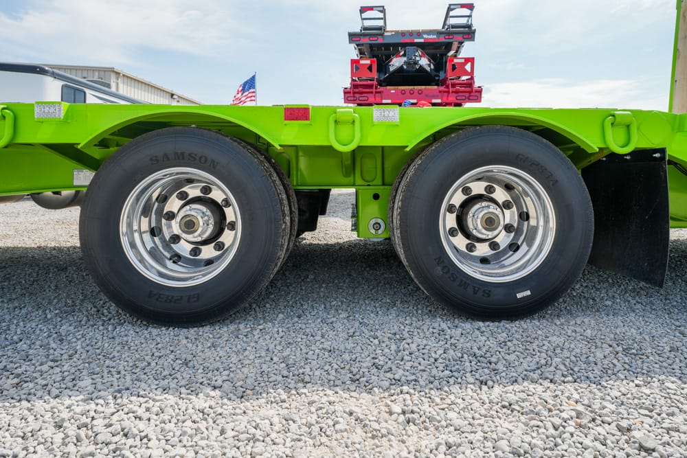 Close-up of a bright green trailer chassis with two large chrome-rimmed tires on a gravel lot, under a blue sky with equipment in the background.
