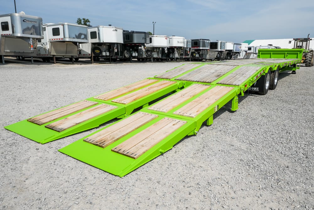 Bright green lowboy trailer with three wooden ramps extended on a gravel lot beside other trailers to load equipment or vehicles.