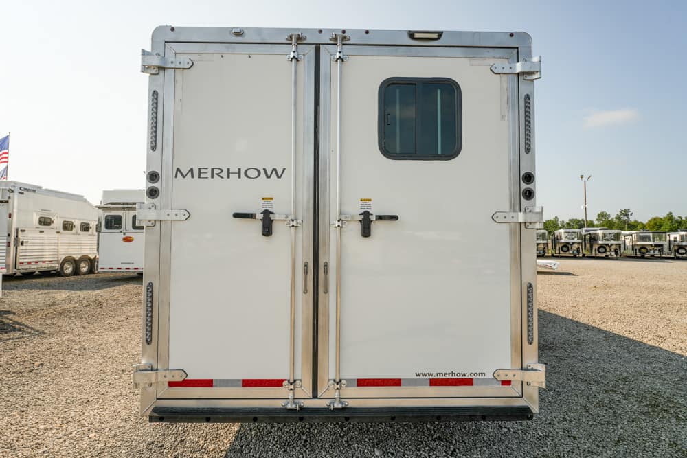 Rear view of a white Merhow horse trailer with dual doors and a small window, parked on a gravel lot.