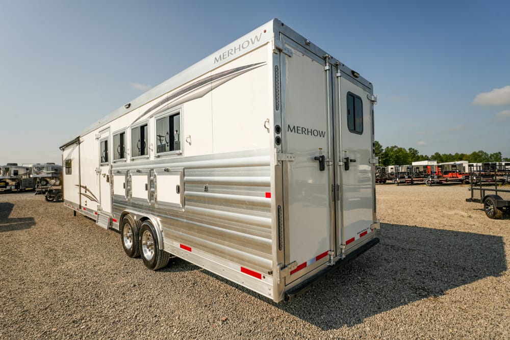 White Merhow horse trailer with aluminum siding and side windows parked on a gravel lot overlooking other trailers in the background