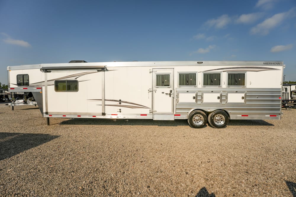 White horse trailer on a gravel lot with multiple windows and storage compartments visible along its side, under a clear blue sky.