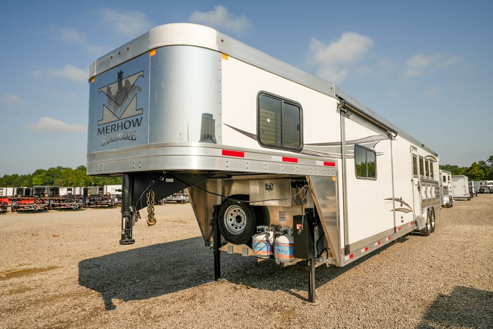 Silver Merhow horse trailer with a raised gooseneck overhang, parked on a gravel lot with propane tanks visible under the front, a spare tire mounted inside the bay area.