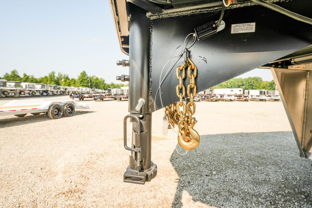 Close-up of a semi-trailer hitch with safety chains and locking latch, in a gravel truck yard with rows of trailers in the background.
