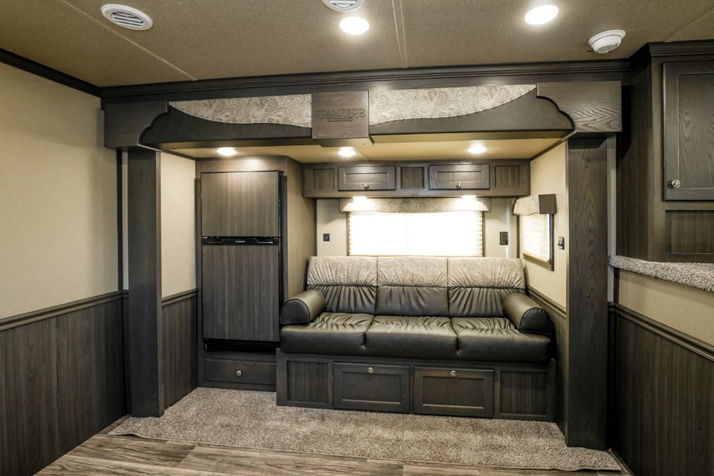 Dark wood RV living area with a black leather sofa, overhead cabinets, fridge on the left, and a window with blinds behind the sofa.