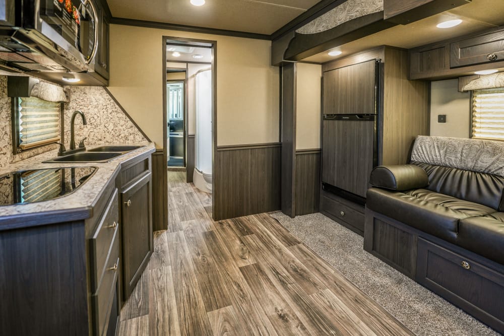Modern RV interior: galley kitchen along left with sink, stove, and dark wood cabinets; black leather sofa on the right with overhead storage.