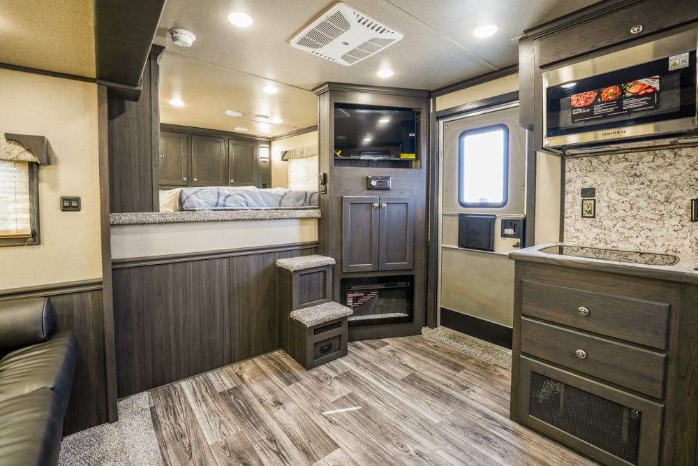 Interior of a modern RV living area with a kitchenette, microwave, and dark wood cabinets along the right, and a sleeping loft above a small step stool area on the left