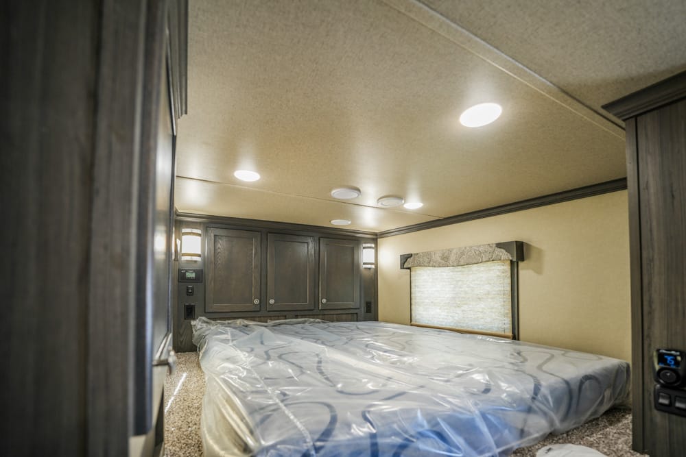 RV bedroom with a large bed wrapped in plastic, dark wood cabinets along the wall, and a window with a shade.