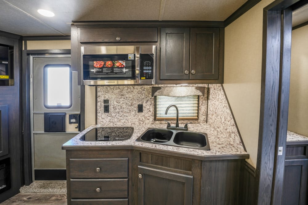 Compact RV kitchen with dark wood cabinets, marble-like countertop, and a double sink beneath a microwave.