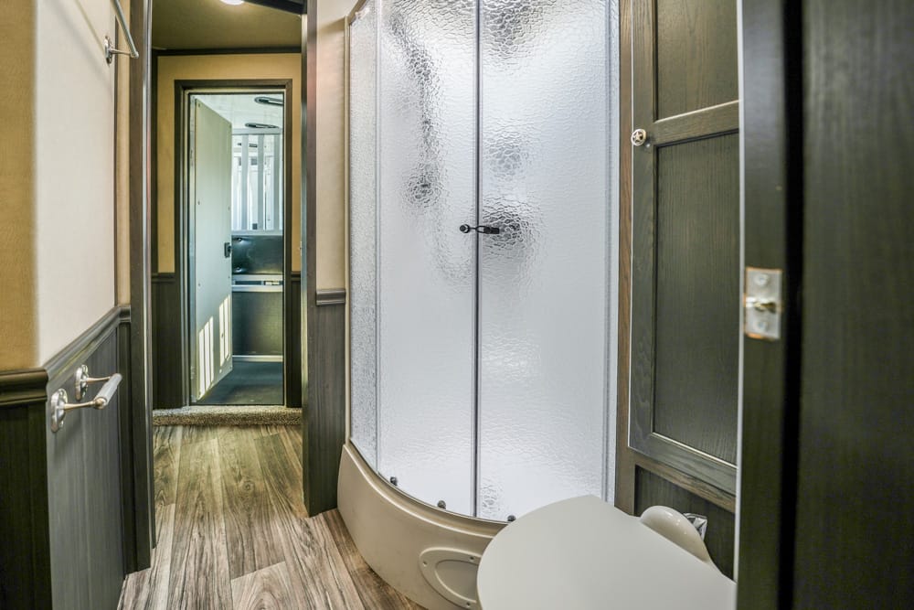 Compact RV bathroom with a curved frosted-glass shower enclosure, toilet in foreground, and dark wood cabinets along a wood-look floor | Small hallway leads to another room visible through an open door.
