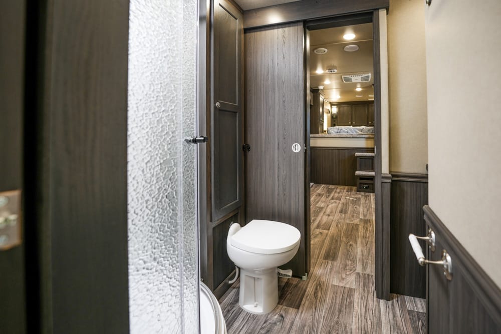 Small RV bathroom with white toilet and dark wood paneling, open doorway leading to a bedroom area with wood floors and cabinetry