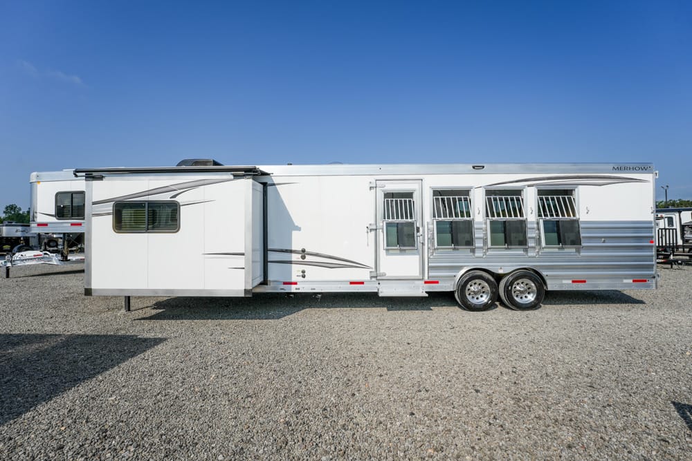 Long white horse trailer with multiple side windows and dual axles parked on a gravel lot under a clear blue sky.