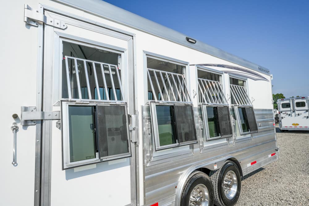 White aluminum horse trailer with multiple large windows covered by metal bars, parked on a gravel lot with another trailer in the background.