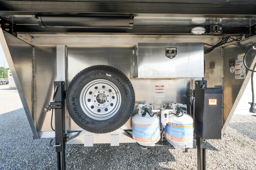 Underbelly of a utility trailer with a mounted spare tire, two propane cylinders, and metal storage boxes.