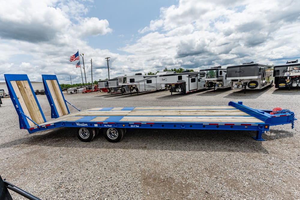 Long blue flatbed trailer with wooden deck and two ramps at the rear, parked on a gravel lot with campers in the background.