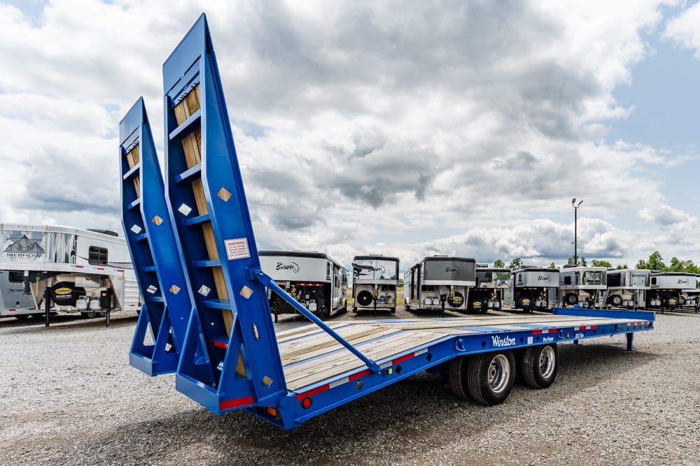 Blue flatbed trailer with raised loading ramps in a gravel lot, surrounded by other trailers and RVs.