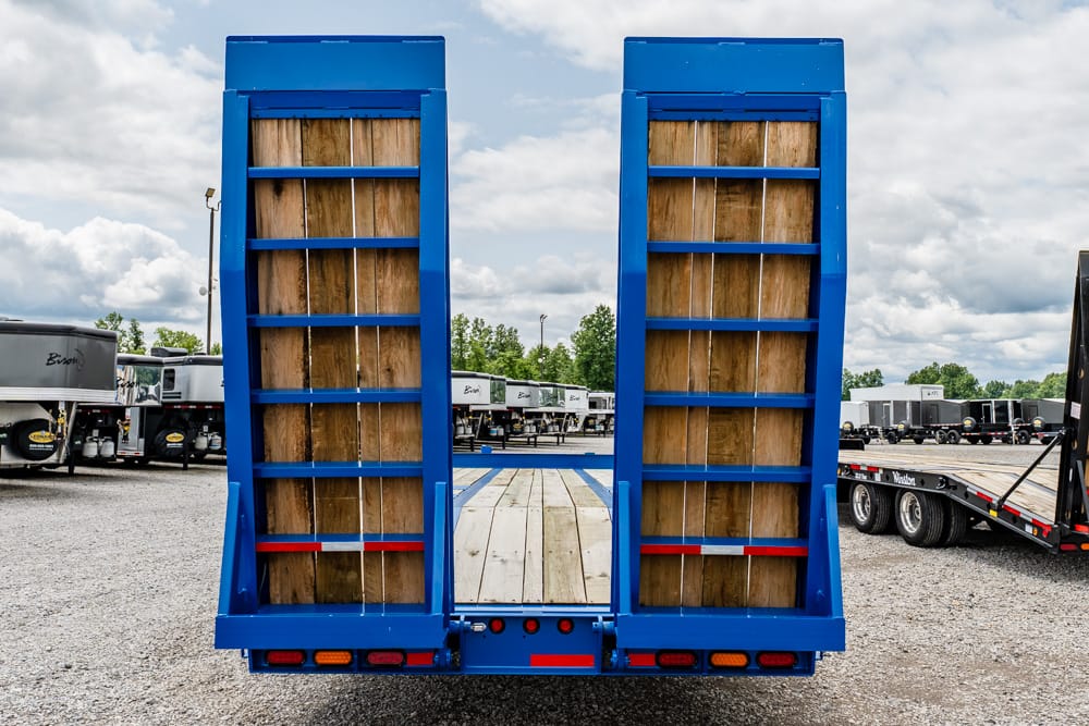 Back view of a blue truck trailer with tall wooden loading ramps ready for loading vehicles or equipment in a lot.