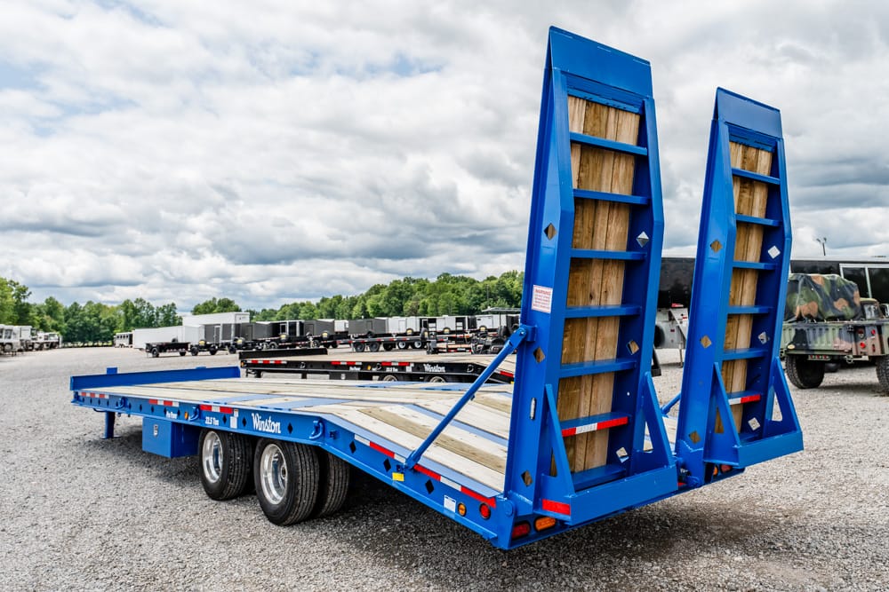 Blue flatbed trailer with fold-down loading ramps raised, parked on a gravel lot at a trucking yard