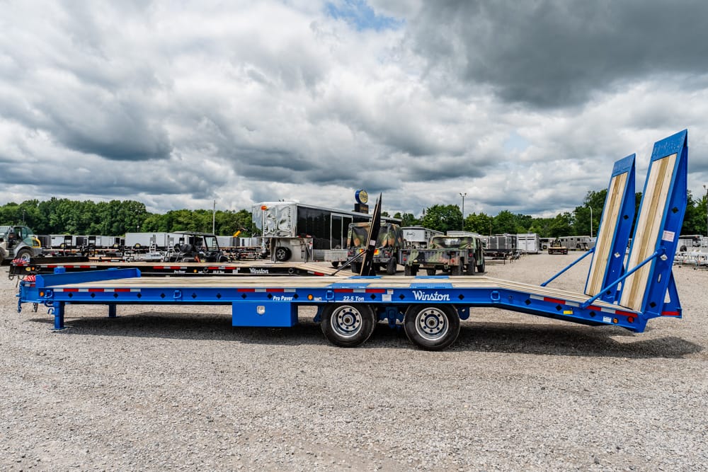 Blue Winston car hauler trailer with raised loading ramps in a gravel lot, surrounded by trucks and trailers in the yard.