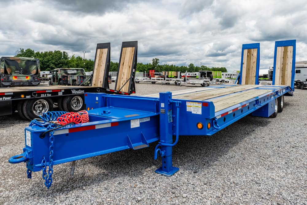 Blue lowboy trailer with wooden deck and rear ramps raised, parked on a gravel lot for heavy equipment transport.