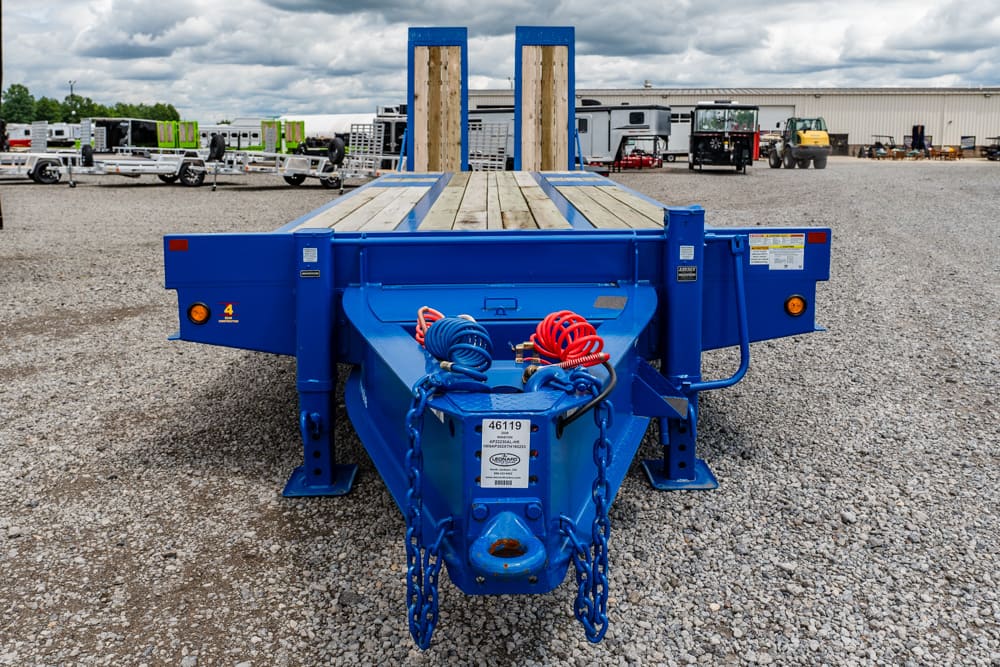 Blue heavy-duty flatbed trailer in a gravel lot, with wooden deck and rear hitch chains visible