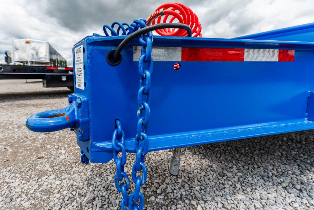 Blue utility trailer tongue with heavy blue chain and a red coiled cord resting on a gravel surface underneath a cloudy sky.