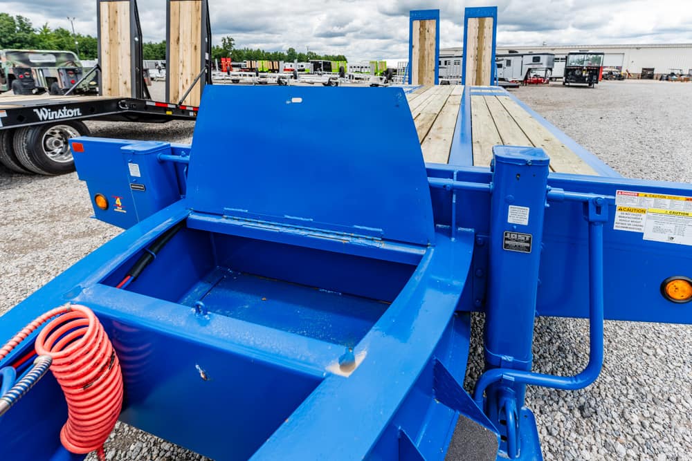 Blue heavy‑duty trailer with a wooden deck, parked on a gravel lot; a red coiled hose sits in the foreground and safety labels are visible.