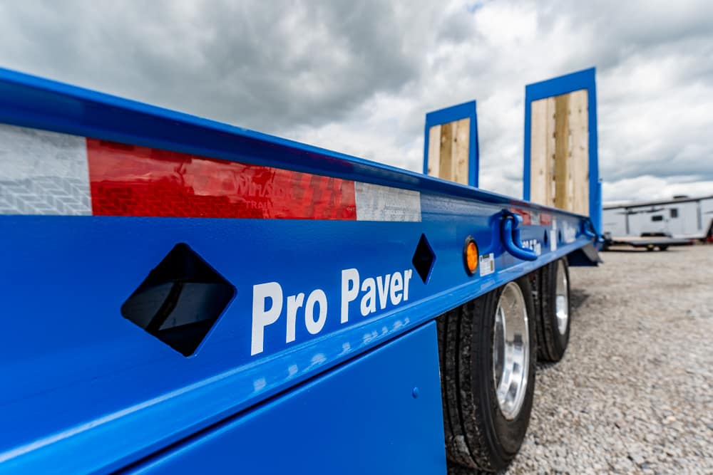 Blue Pro Paver flatbed trailer with dual axles parked on a gravel lot, logo visible along the side.