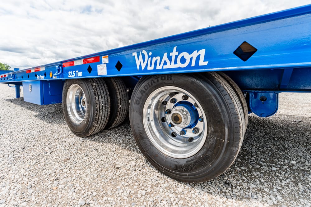 Blue Winston flatbed trailer with dual-axle wheels on a gravel lot, text 'Winston' and '22.5 Ton' along the side visible.