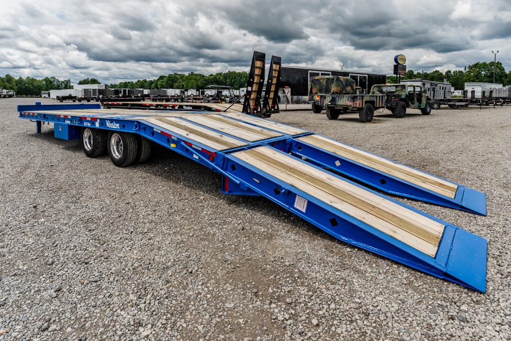 Blue lowboy trailer with wooden ramps parked on a gravel lot, ready for loading heavy equipment in the background.