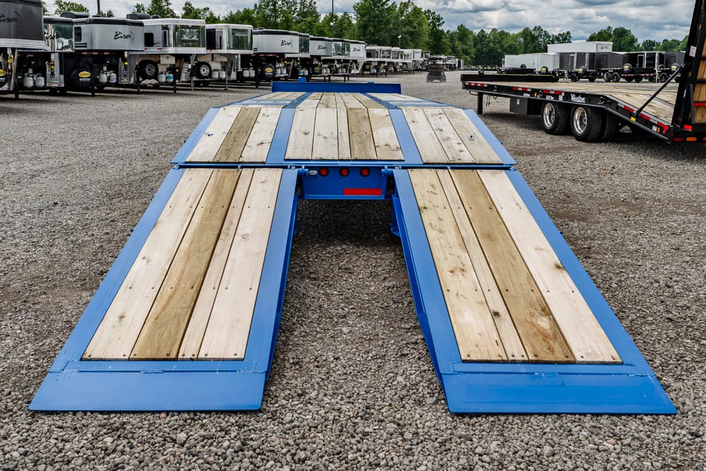 Blue industrial loading ramp with wooden planks in a gravel trailer yard; used for tractor-trailers behind.
