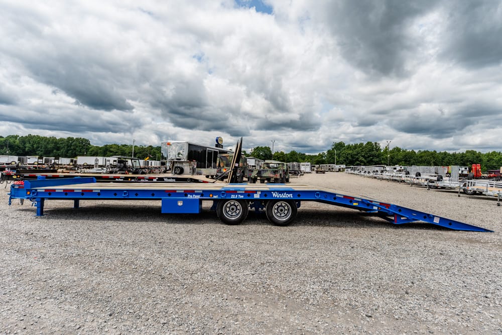 Blue low-bed trailer with a ramp extended in a gravel lot filled with trailers and equipment in the background.