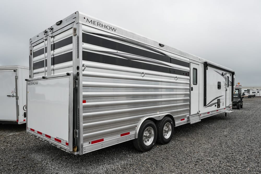 Two-axle silver Merhow horse trailer parked on a gravel lot, attached to a truck in the distance.