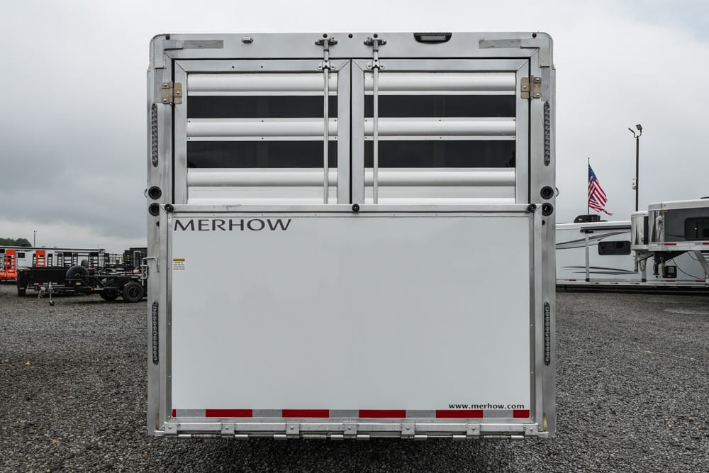 Rear view of a silver Merhow horse trailer with vented doors, parked on a gravel lot; an American flag is visible in the background.