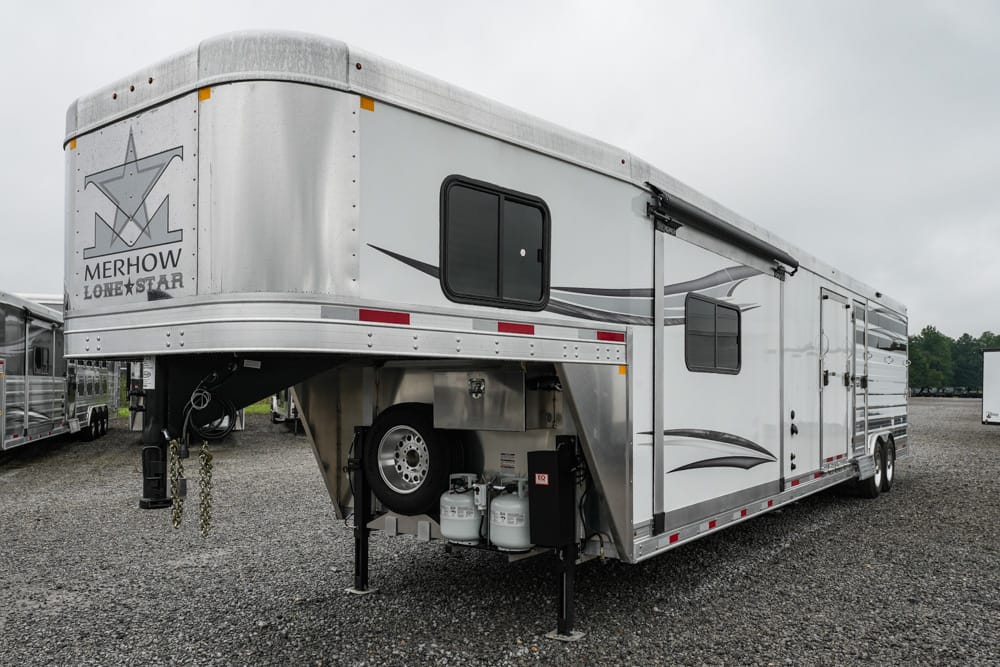 Silver Merhow Lone Star gooseneck horse trailer on a gravel lot, raised front section with two windows and propane tanks beneath.
