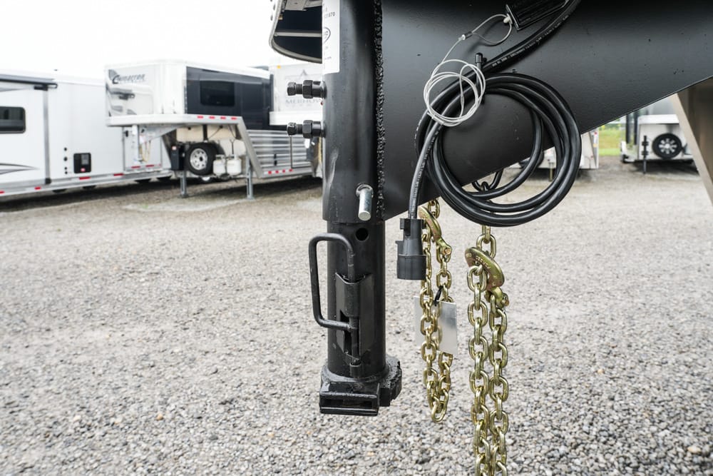 Close-up of a trailer hitch with safety chains and coiled electrical cable on a gravel lot, with other trailers in the background.