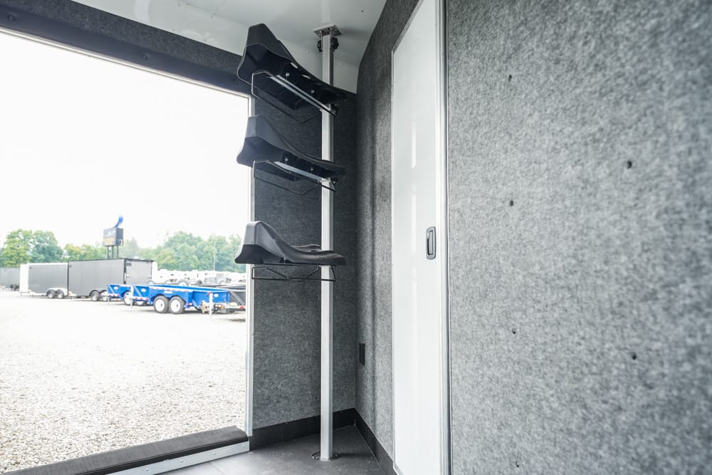 Interior of a trailer with a vertical rack holding three black helmets on metal supports in a grey-walled space.