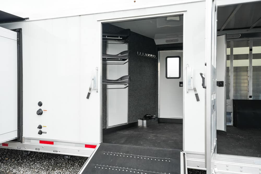 Open white trailer with a storage area inside, featuring wall-mounted racks, hooks, and a small back doorway visible at the far end.
