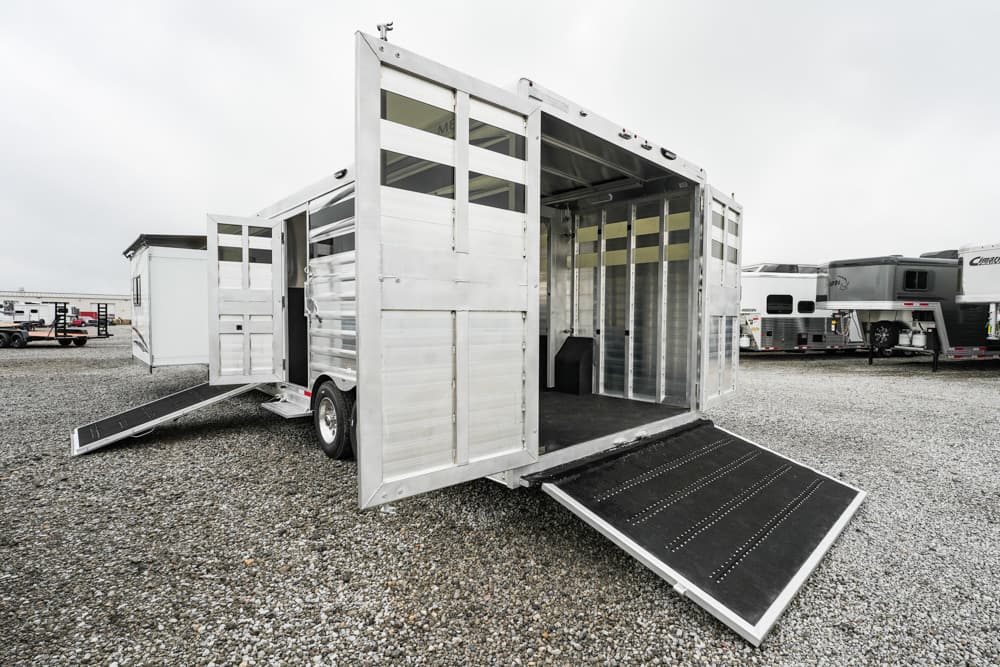 Silver aluminum horse trailer with the rear ramp down and front open doors in a gravel lot; other trailers visible in the background