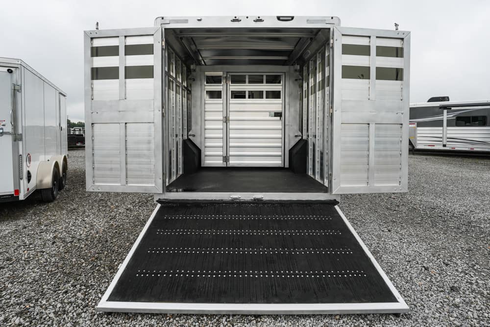 Aluminum livestock trailer with a lowered loading ramp on a gravel lot, showing an empty interior with partitions and a rubber floor edge.