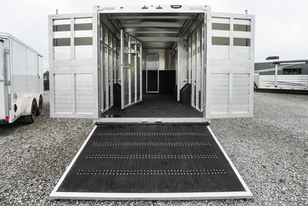 Aluminum horse trailer with ramp down on a gravel lot, interior stalls visible.