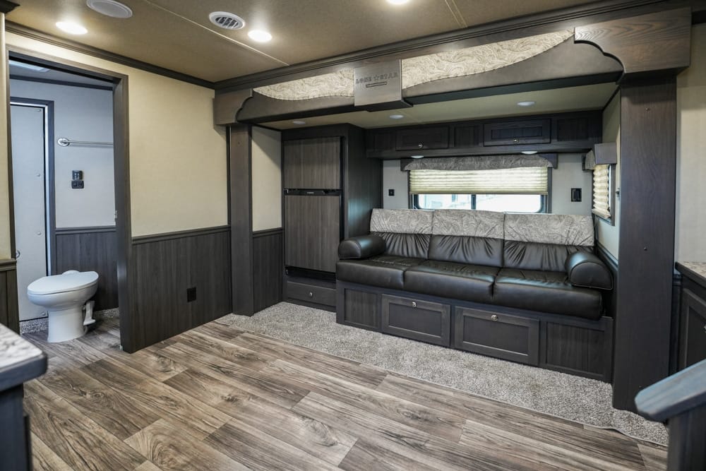Interior of a modern RV living area with a dark wood entertainment wall, black leather sofa, and built-in storage under the seating along one wall; a window with shade behind the sofa and overhead cabinets above.