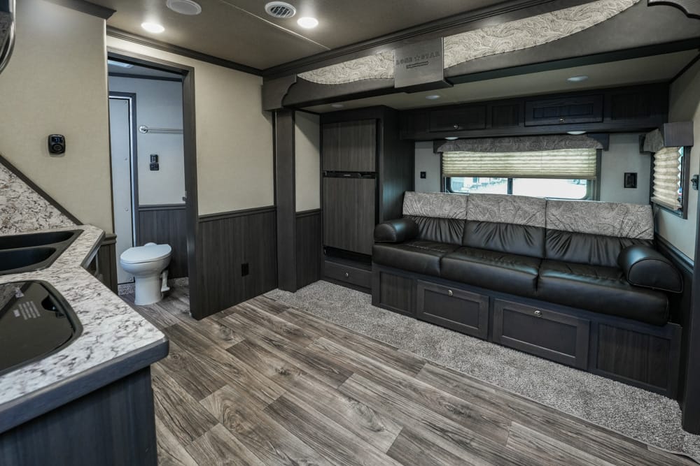Interior of a modern RV: dark wood cabinets, a long black leather sofa with hidden storage, and a kitchen counter with a sink on the left.