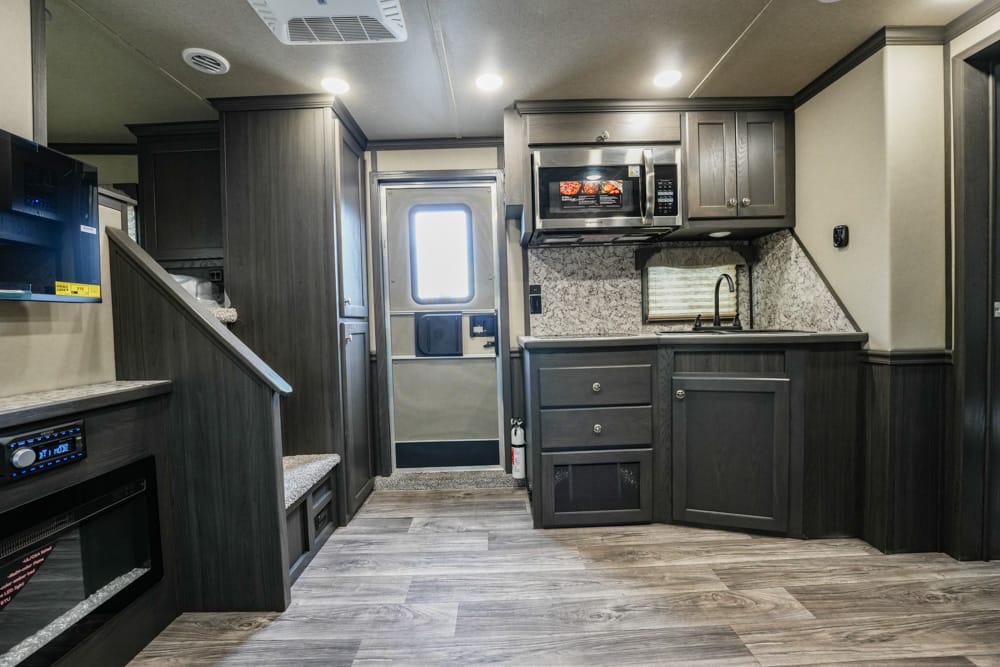 Dark wood RV kitchen with a marble backsplash, microwave, sink, and ample cabinetry surrounding a tiled countertop, plus a doorway to the outside at the rear center of the image.