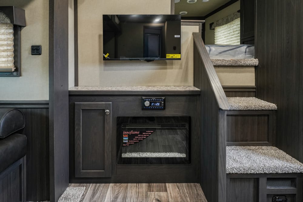 Wall-mounted TV above an electric fireplace in a dark-wood RV living area, with carpeted stairs to the right.