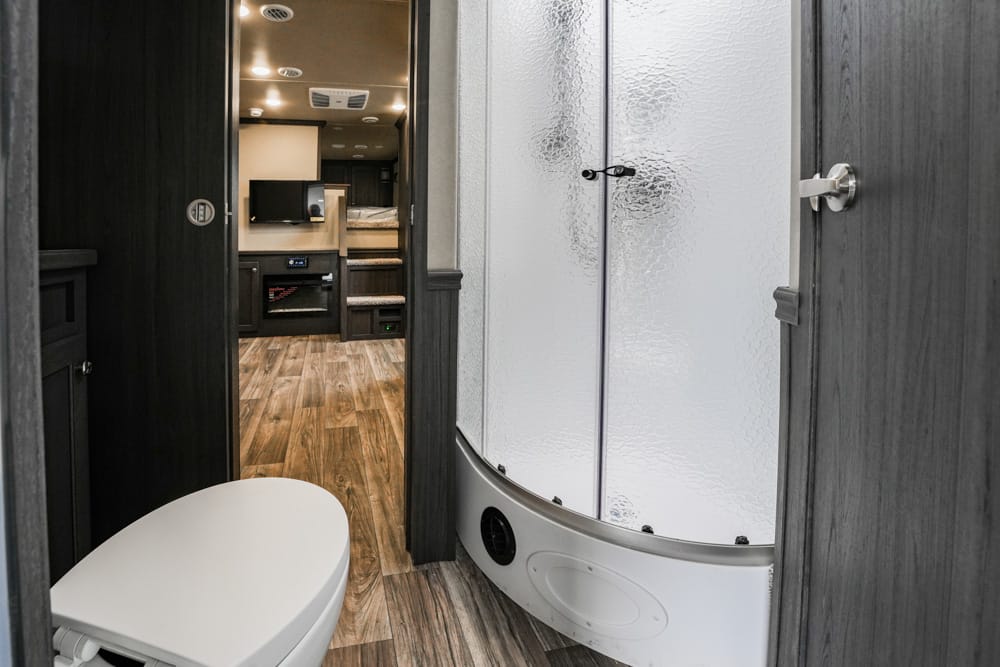 Compact RV bathroom with curved frosted-shower enclosure, toilet, and wood‑grain flooring; doorway to living area visible in the background.